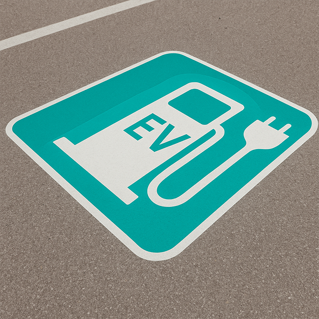 Zdjęcia miejsca parkingowego, a na nim znak poziomy symbolizujący ładowarkę dla auta elektrycznego z napisem EV w kolorze białym na turkusowym tle.